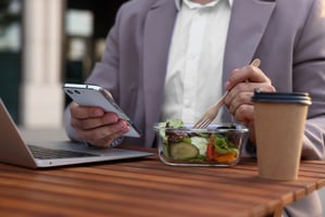 Employee using mobile phone at desk while eating salad lunch and drinking coffee