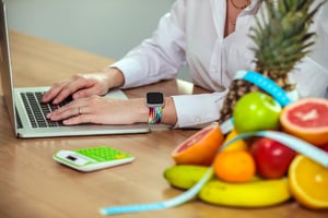 Employee wearing fitness watch, sitting at office desk next to a bowl of healthy fruits