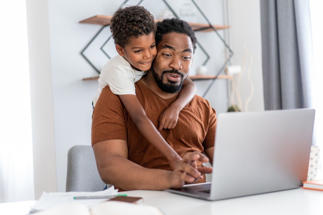Playful child climbing on his father's shoulders while he works in home office